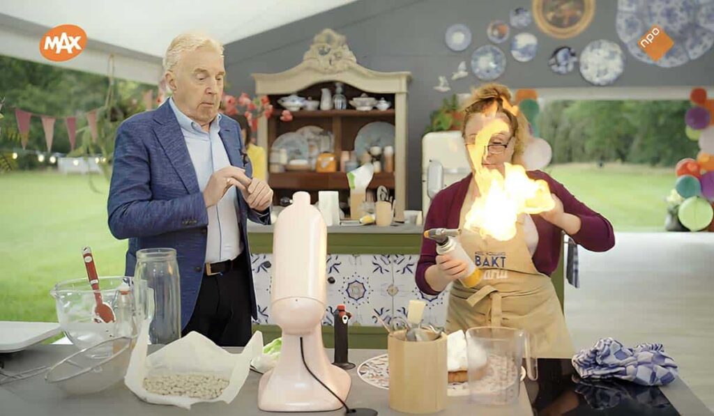 Heel Holland Bakt 2024 presentator André van Duin en deelneemster Carla staan bij een keuken in de tent en de vrouw heeft een brander met steekvlam vast